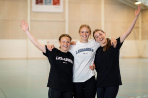 3 piger fra Lægården Idrætsefterskole står i deres sportshal mens de har armene i vejret og smiler
